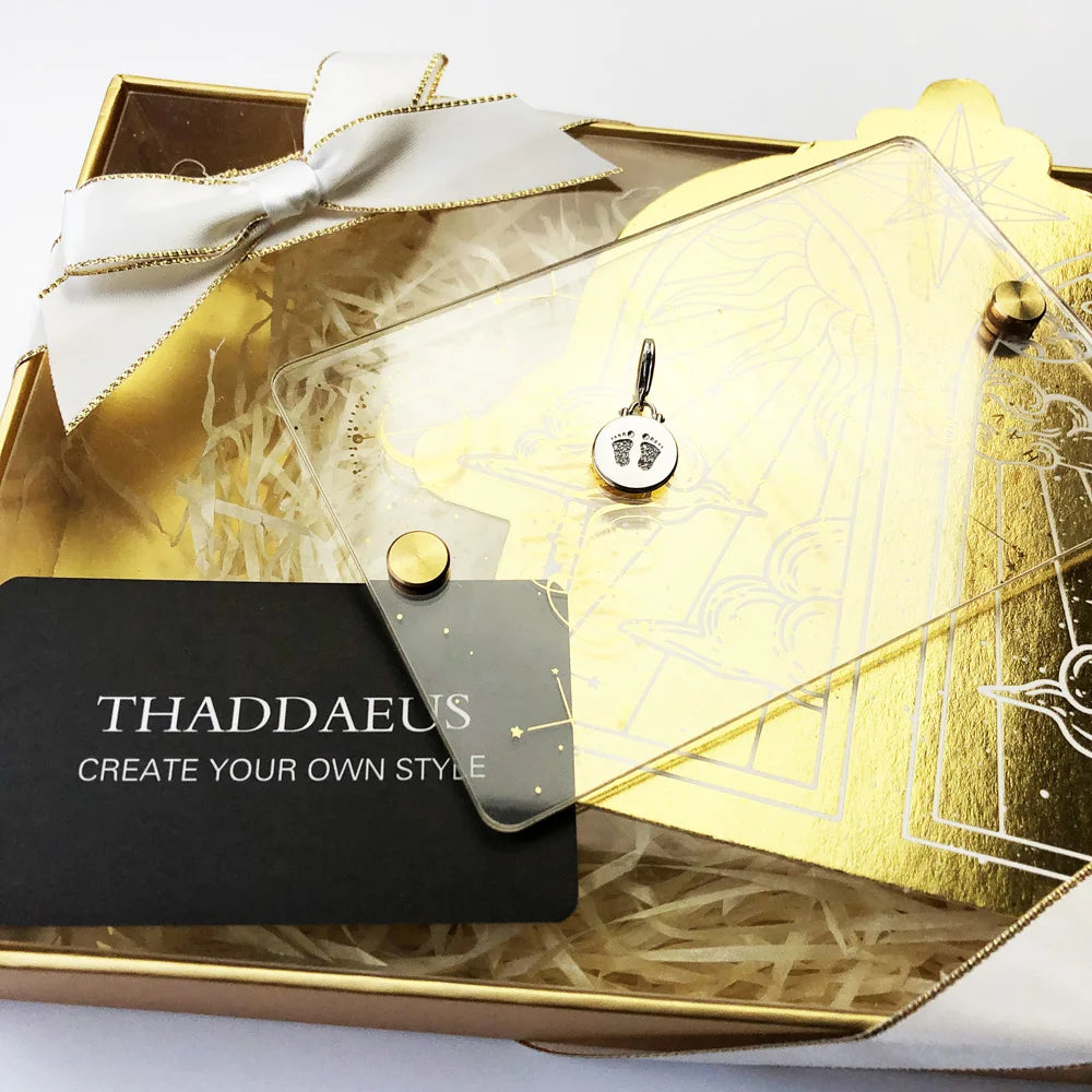 Silberner Anhänger mit runder Disc und Baby-Fußabdruck auf goldfarbener Geschenkverpackung.