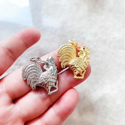 Hahn-Anhänger in silberfarben und goldfarben in einer Hand gehalten