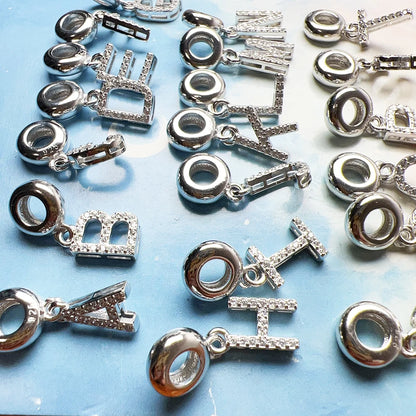 Silberne Charm-Anhänger in Buchstabenform mit hellem Steinbesatz, mehrere Motive